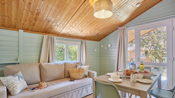 A cozy room with a wooden ceiling, a light green wall, a sofa with cushions, a table with breakfast items, and a view of greenery outside.