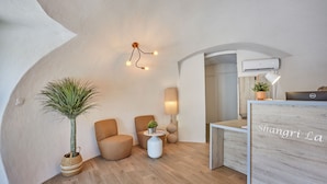 A modern room with a wooden floor, a potted plant, two armchairs, a small table with a lamp, and a desk with a computer.