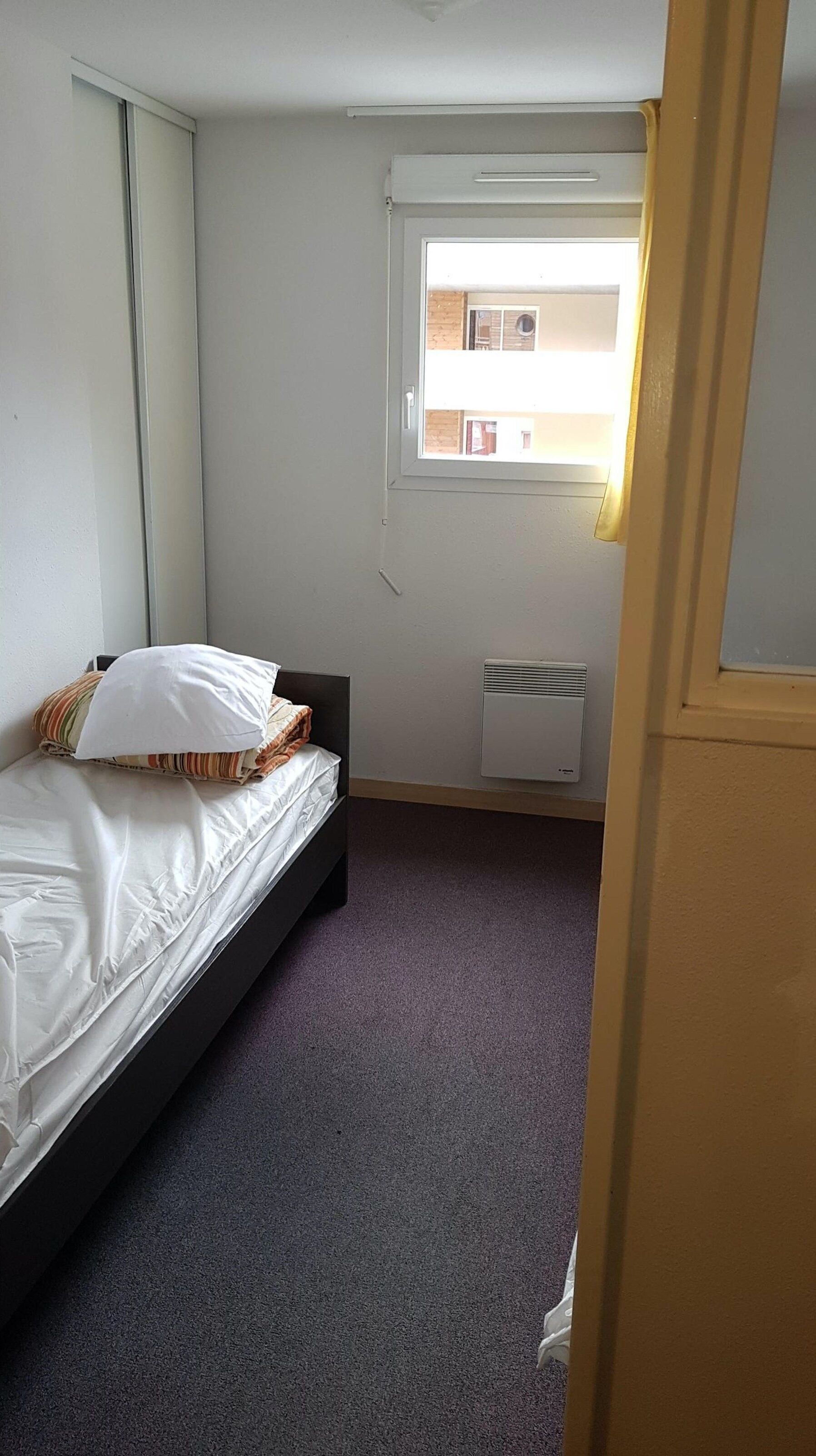 A small, single-bed room with a window, a radiator, and a door.