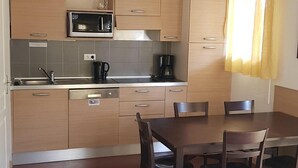 A compact kitchen with a microwave, coffee maker, and dishwasher. There is a dining table with four chairs and a sofa in the foreground.