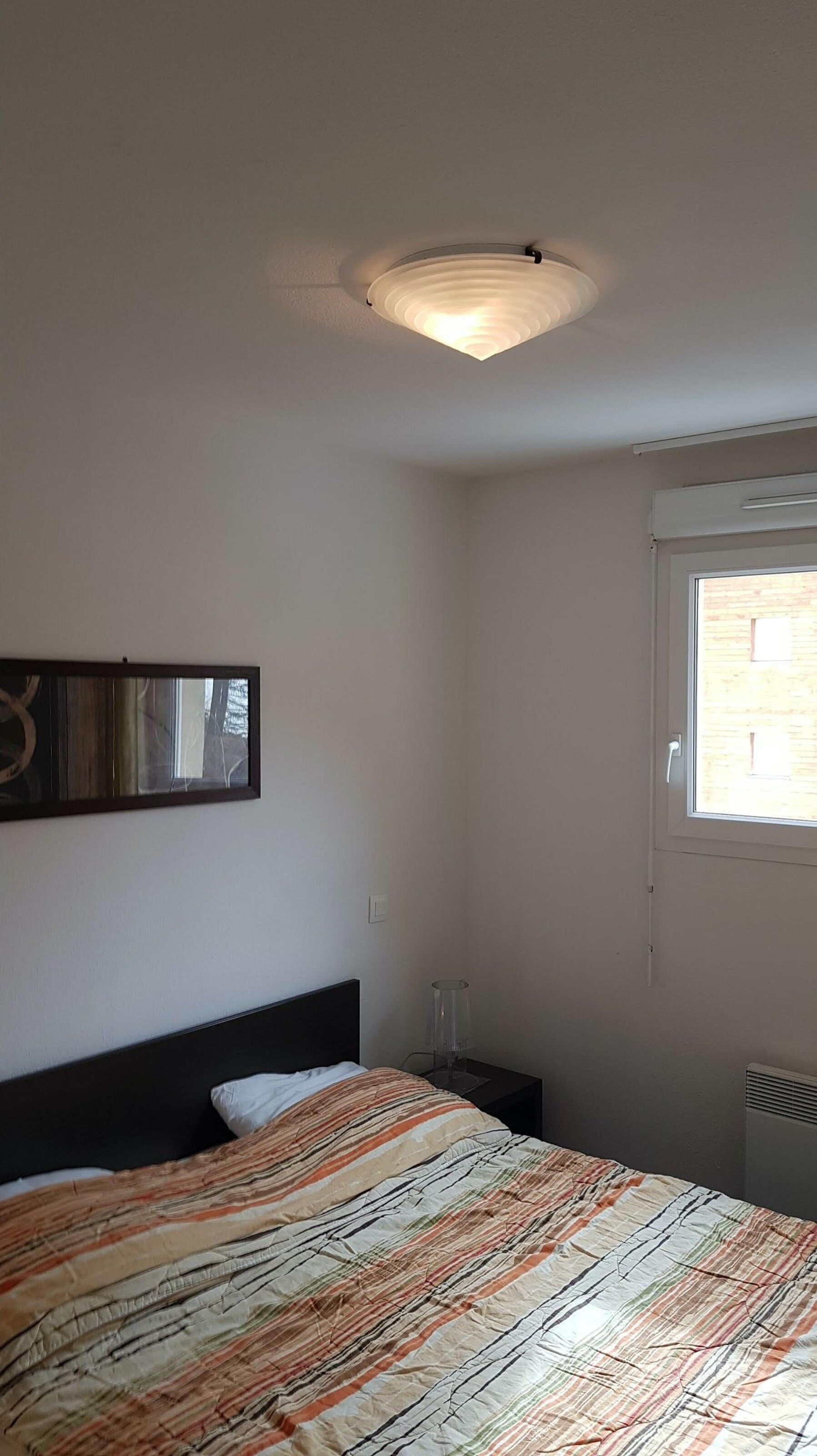 Ein Schlafzimmer mit Bett, Nachttisch, Lampe, Deckenleuchte, Fenster und einem Wandbild.