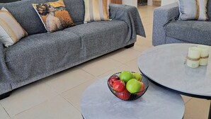 A modern living room with grey sofas, a round coffee table with fruit, and a dining area with a chandelier. - Serenity Pearl villa with a private pool,  2 min walk to the beach (Protaras)
