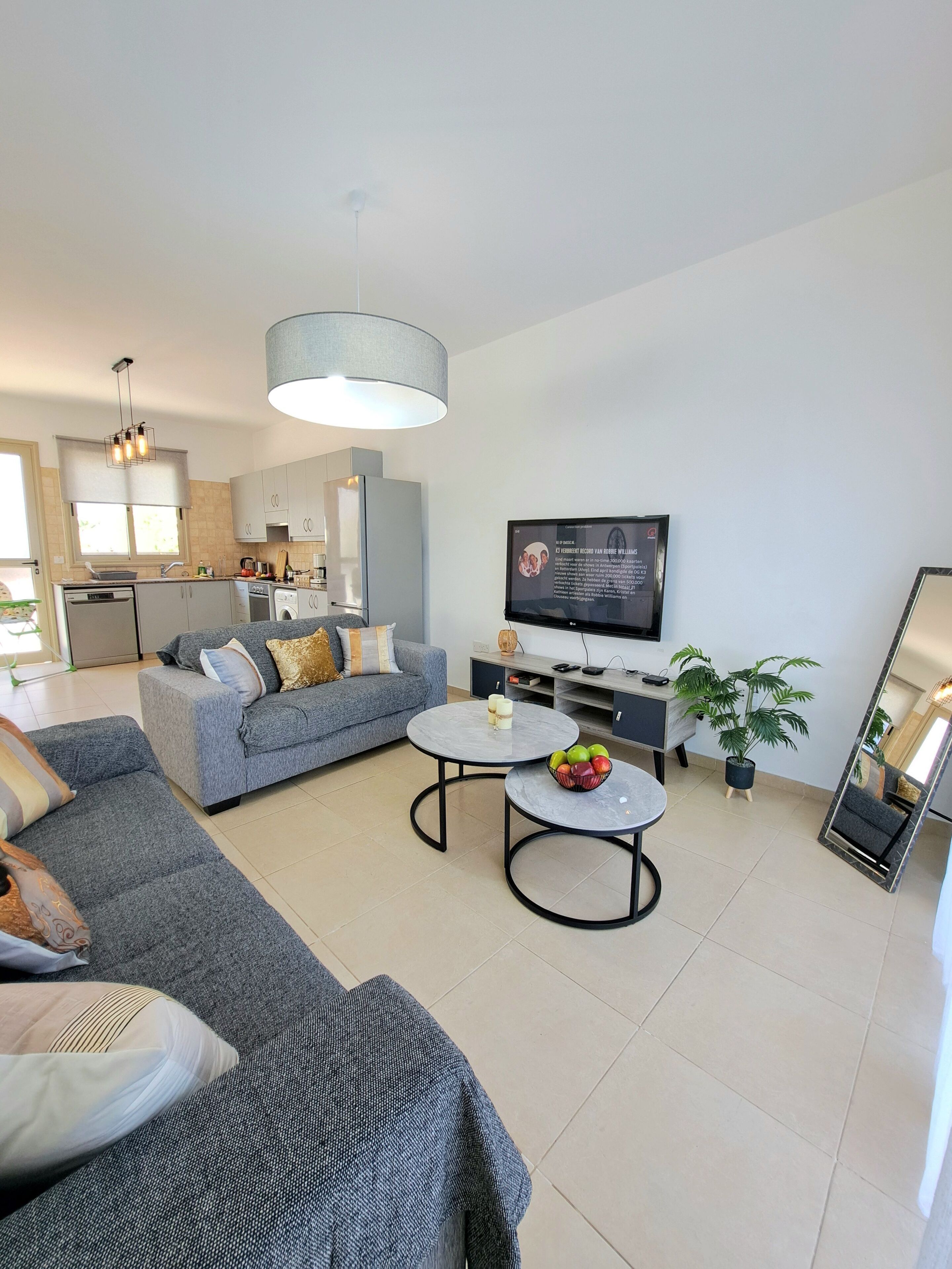 A modern living room with a grey sofa, two round coffee tables, a flat-screen TV, and a kitchen in the background.