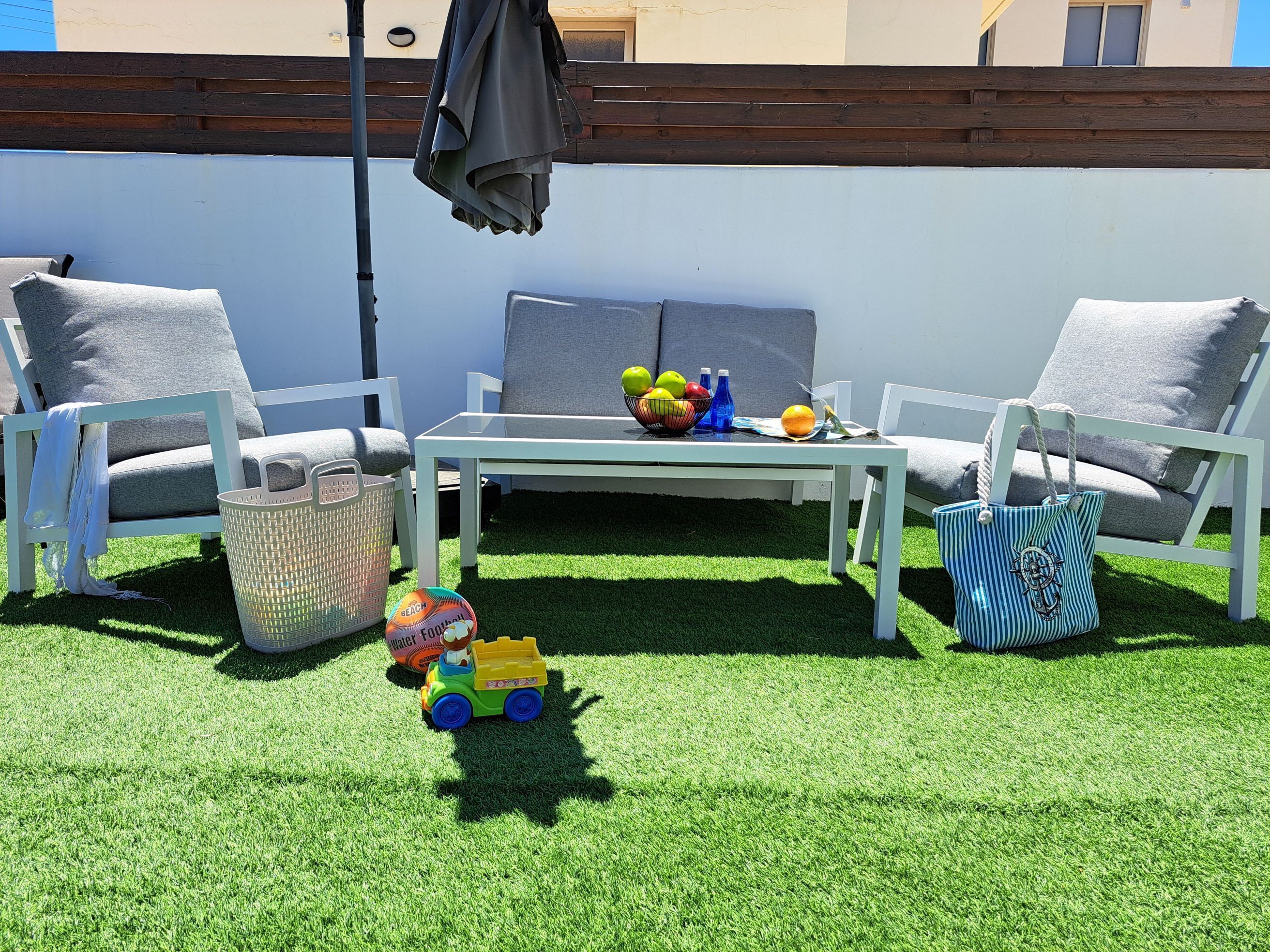 Eine Terrasse mit künstlichem Rasen, zwei weiße Stühle, ein Tisch, ein Korb, ein Spielzeugauto und eine Tasche.