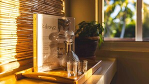 Una habitación con ventana, persiana de bambú, un decantador de vidrio y una bandeja de vidrio con vasos pequeños.