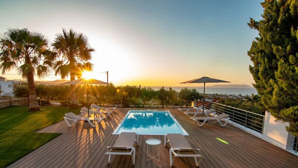Eine Holzterrasse mit Pool, Liegestühlen und Blick auf das Meer bei Sonnenuntergang.