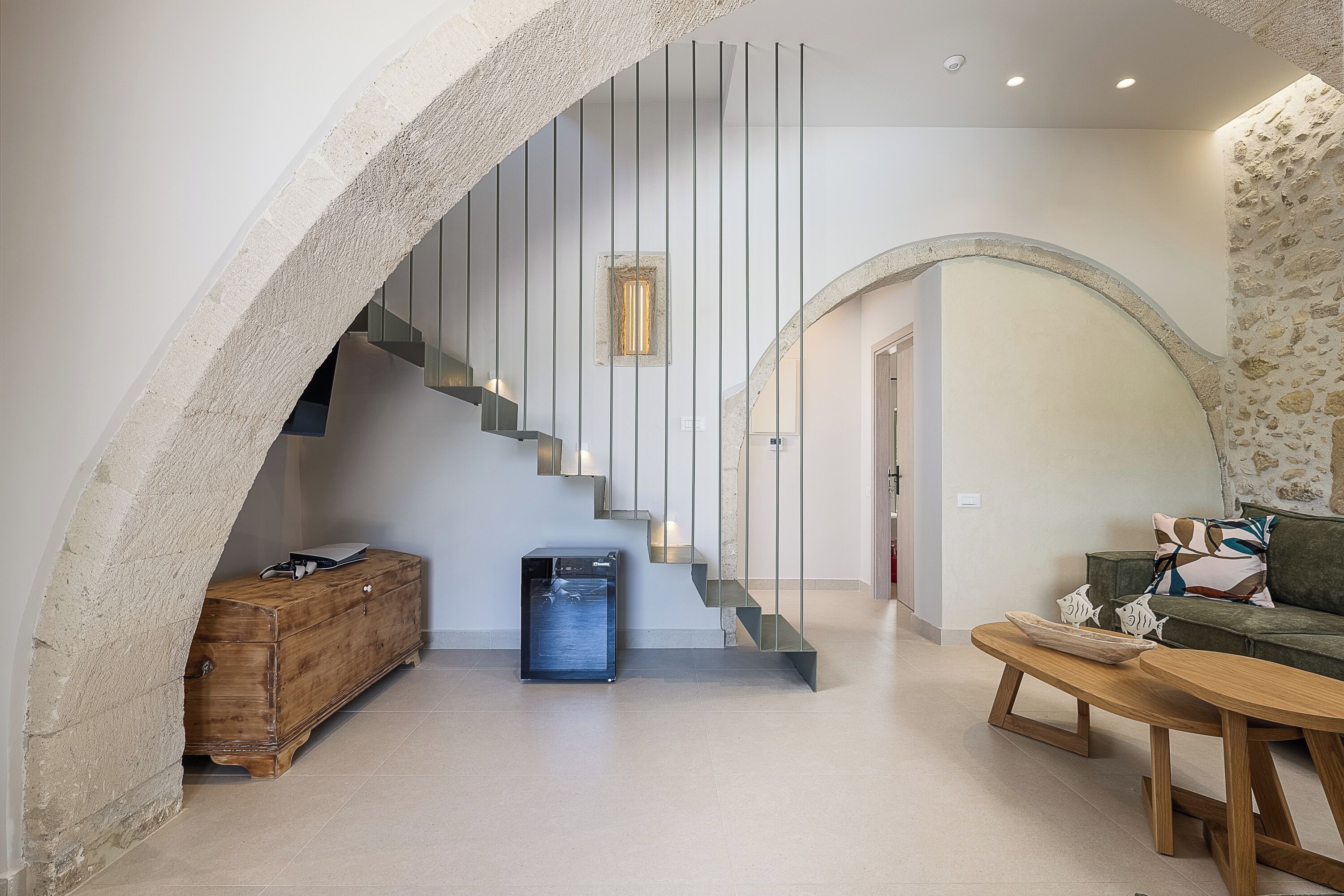 Een modern interieur met een stenen boog, een wenteltrap met verticale lijnen, een houten kist, een blauwe prullenbak en een groene bank met decoratieve kussens.