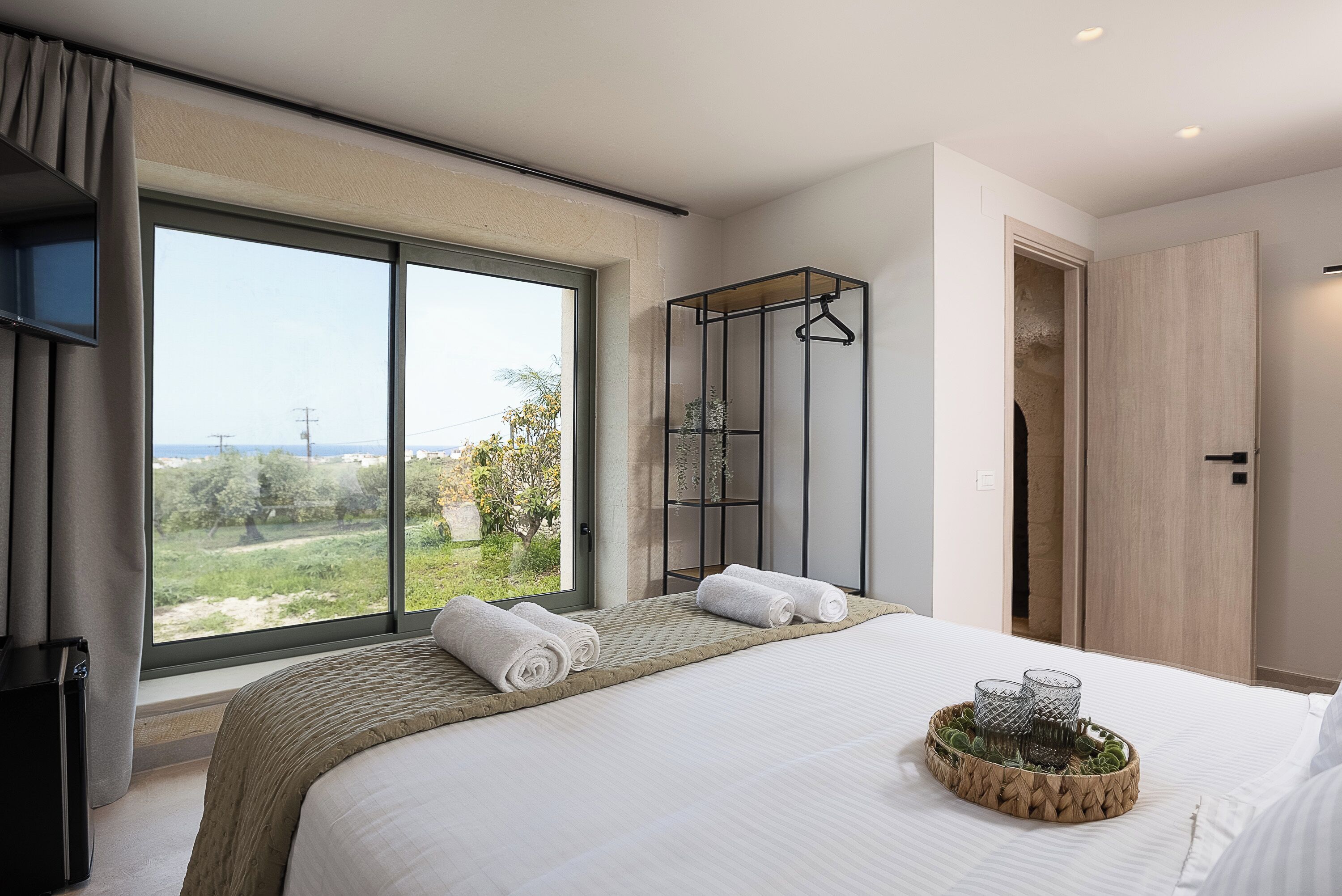 Una habitación de hotel moderna con un ventanal grande, una cama con toallas ordenadas, una mesita de noche con vasos y un armario de madera.