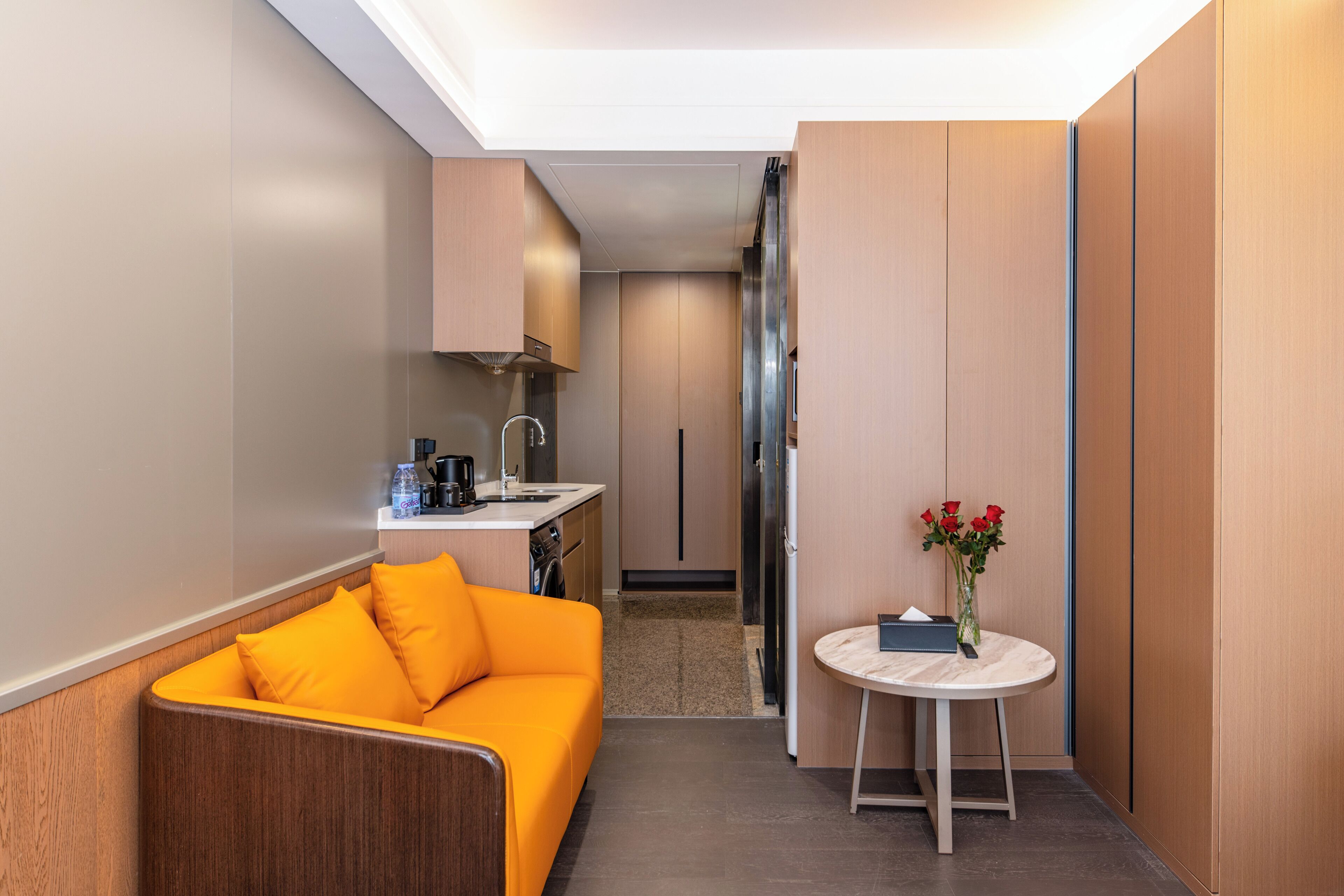Habitación de hotel compacta con kitchenette, sofá, una pequeña mesa redonda con un florero y vistas al pasillo.