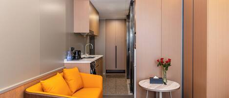 Habitación de hotel compacta con kitchenette, sofá, una pequeña mesa redonda con un florero y vistas al pasillo.