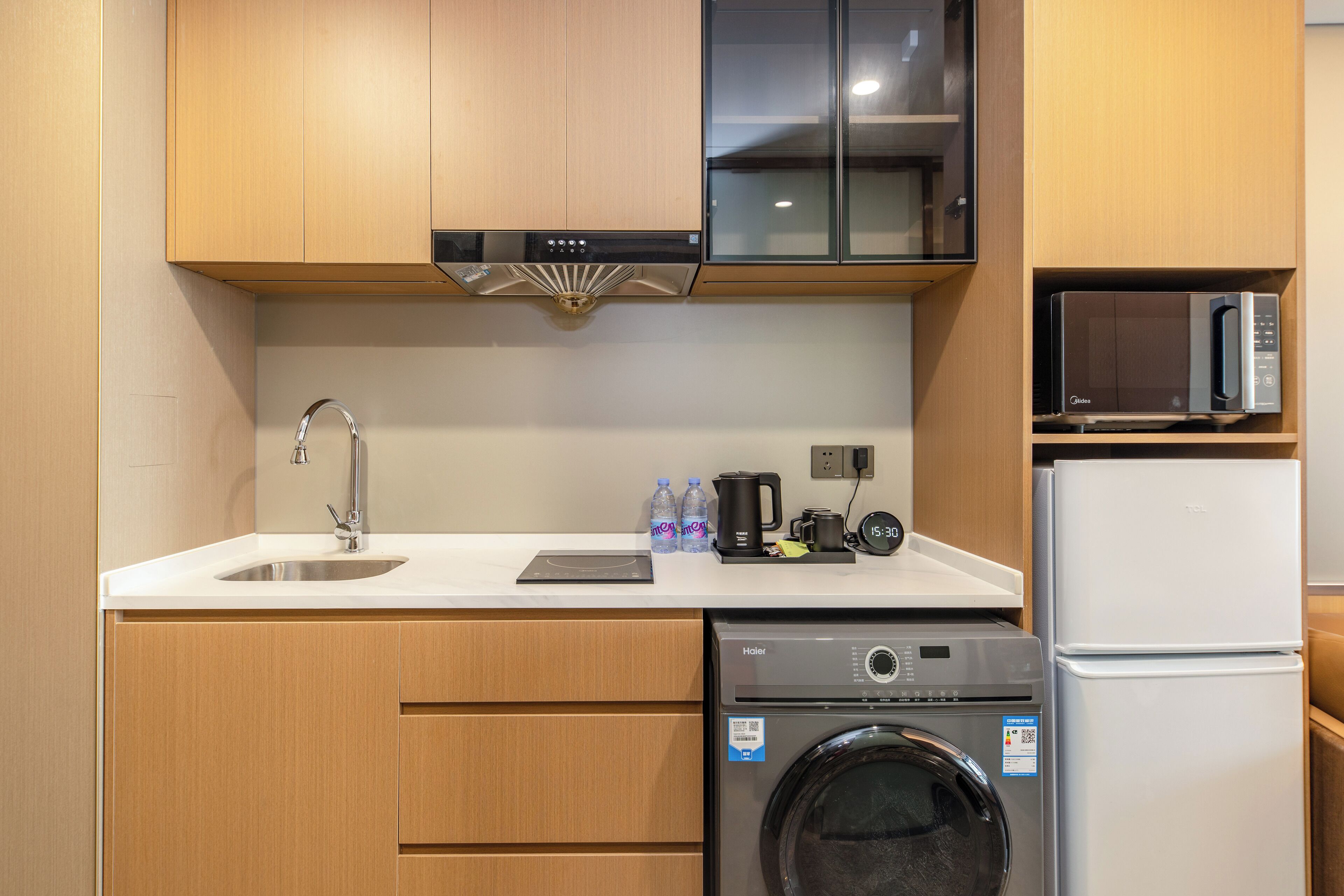 Cocina compacta con armarios de madera, un lavamanos de acero inoxidable, un microondas, un refrigerador y una lavadora.