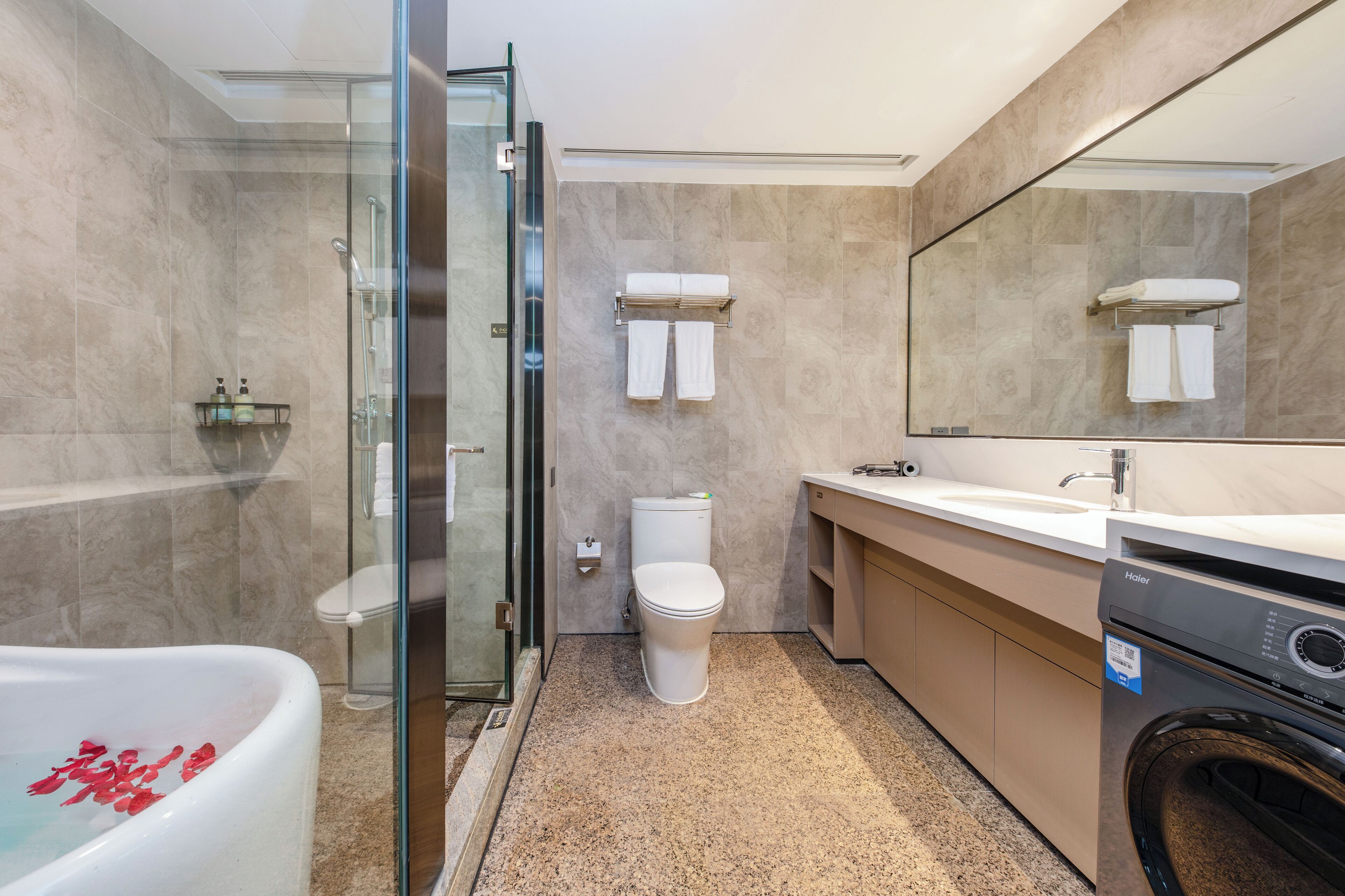 Une salle de bain moderne avec une douche à parois de verre, une baignoire blanche ornée de pétales de roses, des toilettes, un lavabo et un lave-linge.