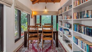A cozy reading nook with a wooden table and chairs, a bookshelf filled with books, and a view of greenery through large windows. - BluSalzz Collection Suvaasa - Ramgarh (Ramgarh)