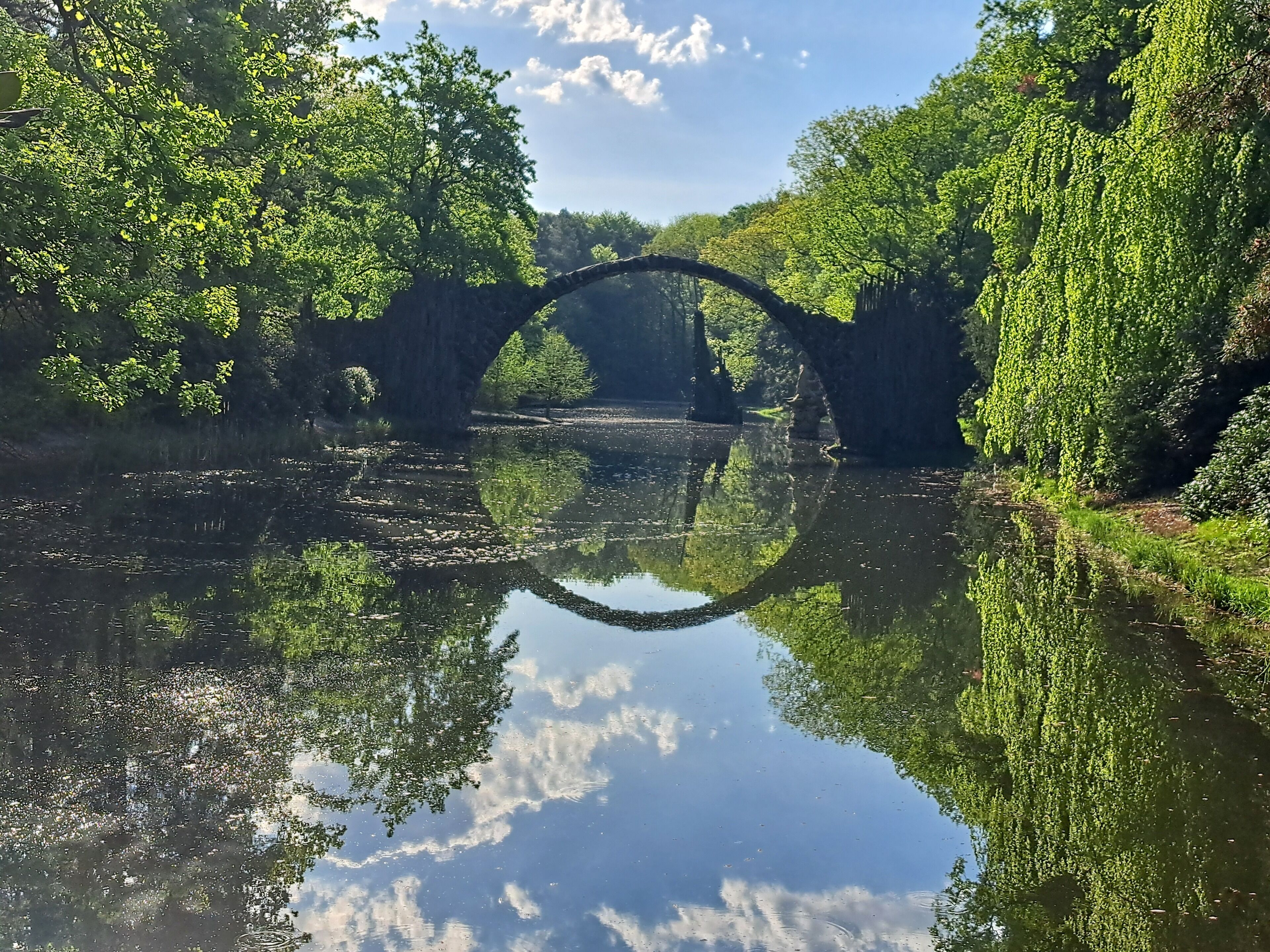 Eine friedliche Flusslandschaft mit einer Steinsbrücke, die sich im Wasser spiegelt, umgeben von üppigem Grün.