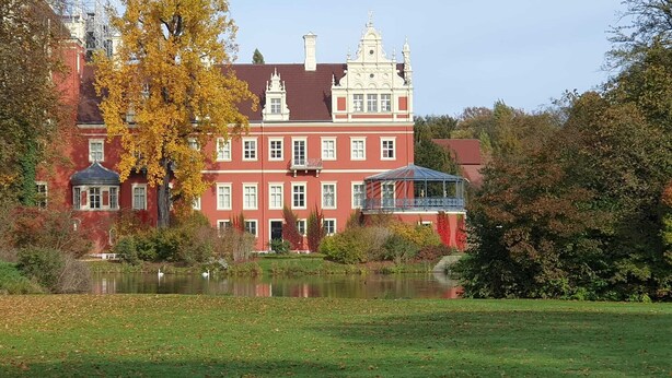 A large red building with multiple windows and a tower, surrounded by trees and a pond. - Parkside Retreat Central location perfect for couples and families (Bad Muskau)