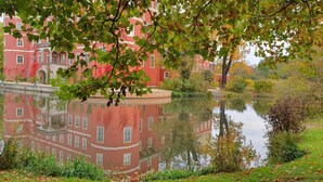 A red building with a reflection in a calm body of water, surrounded by greenery and a tree with autumn leaves. - Parkside Retreat Central location perfect for couples and families (Bad Muskau)