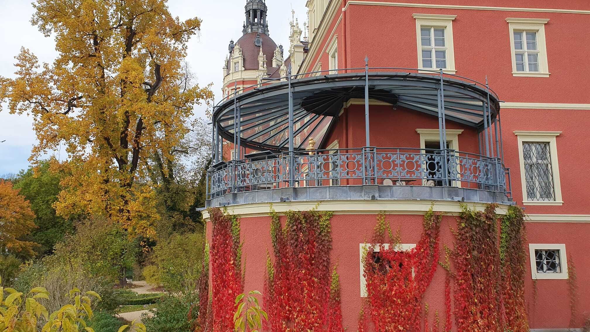 Ein Gebäude mit roter Fassade, einem Balkon mit aufwendiger Metallgitterung und einem Turm mit einer Uhr.