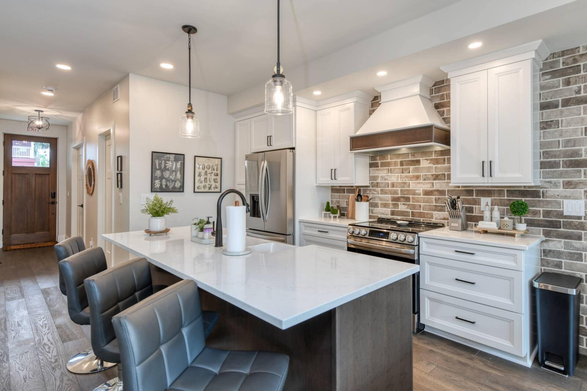 Cocina moderna con armarios blancos, una isla central con asientos, electrodomésticos de acero inoxidable y una pared decorada con ladrillos.