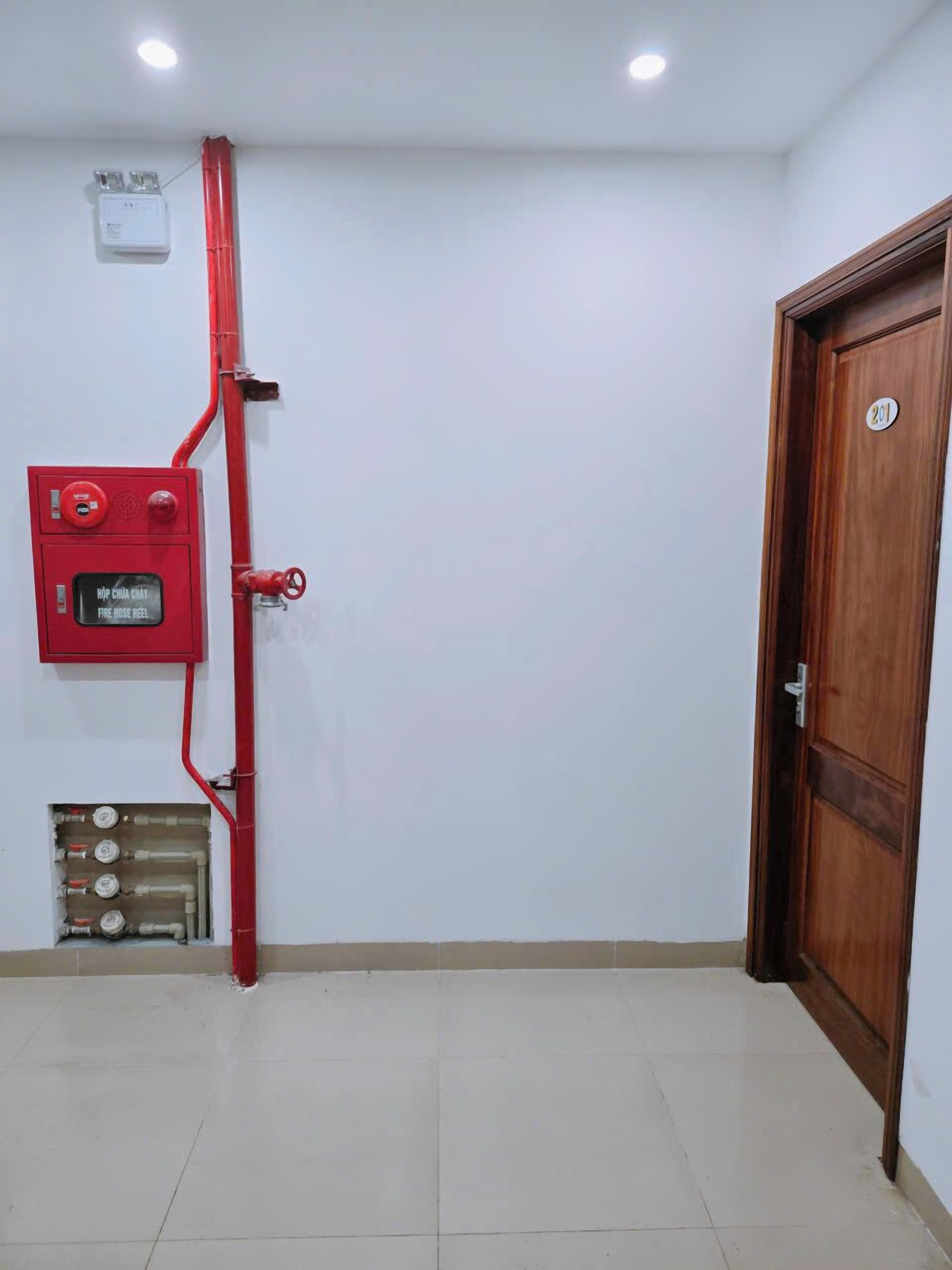 A room with a red fire hose reel, a wooden door with a number plate, and a tiled floor.