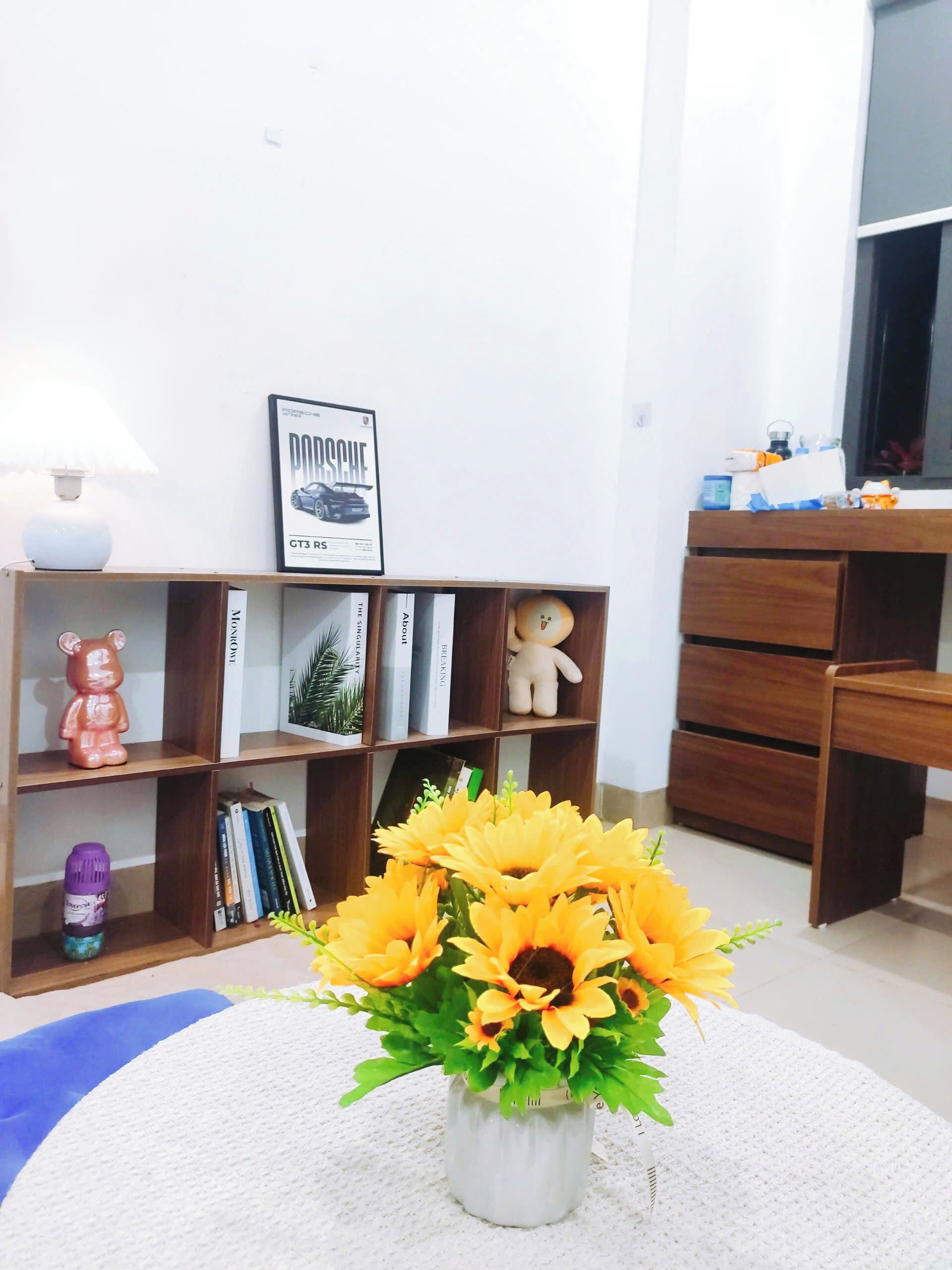 A room with a bookshelf, a framed Porsche GT3 RS poster, a teddy bear, and a vase with sunflowers.