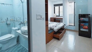 A modern bathroom with a toilet, sink, and shower. There is a bed with a wooden headboard and a small desk with a chair. A vending machine is visible in the corner. - Heart Home (Hanoi)
