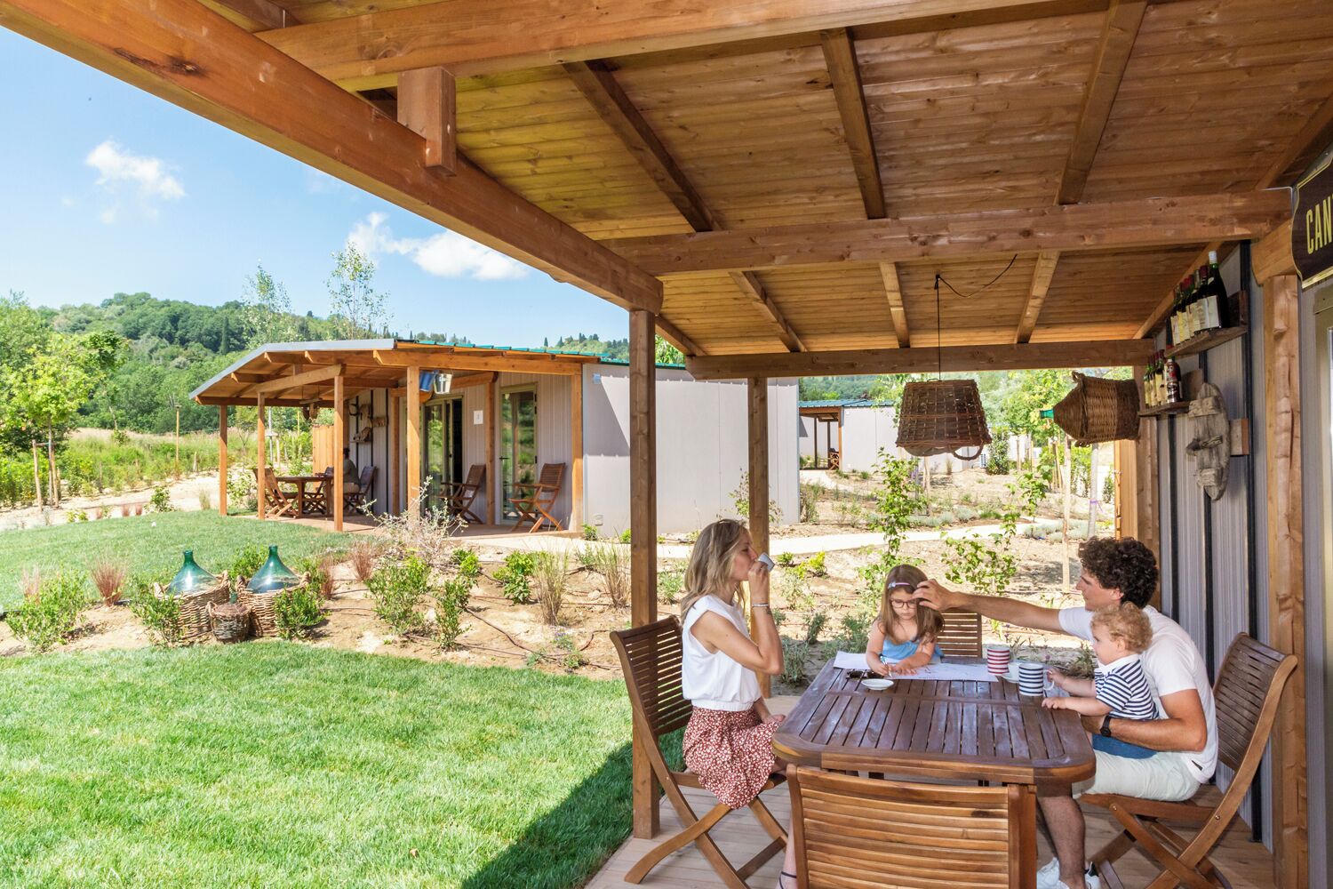 Ein überdachter Außenbereich mit Holztisch und Stühlen, umgeben von einem Garten und einem Gebäude mit dem Schild 'CAN'.