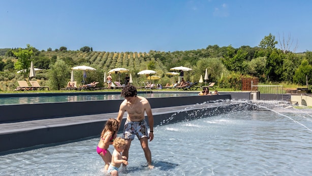 A swimming pool area with lounge chairs and umbrellas, surrounded by a landscaped garden and a hilly backdrop. -  Palagina Il Borgo Agricolo (Figline e Incisa Valdarno)