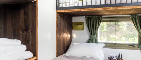 Ein Schlafraum mit Etagenbetten, einem Holzkopfende, einem Nachttisch mit Kamera und Büchern sowie einem Fenster mit Vorhängen.