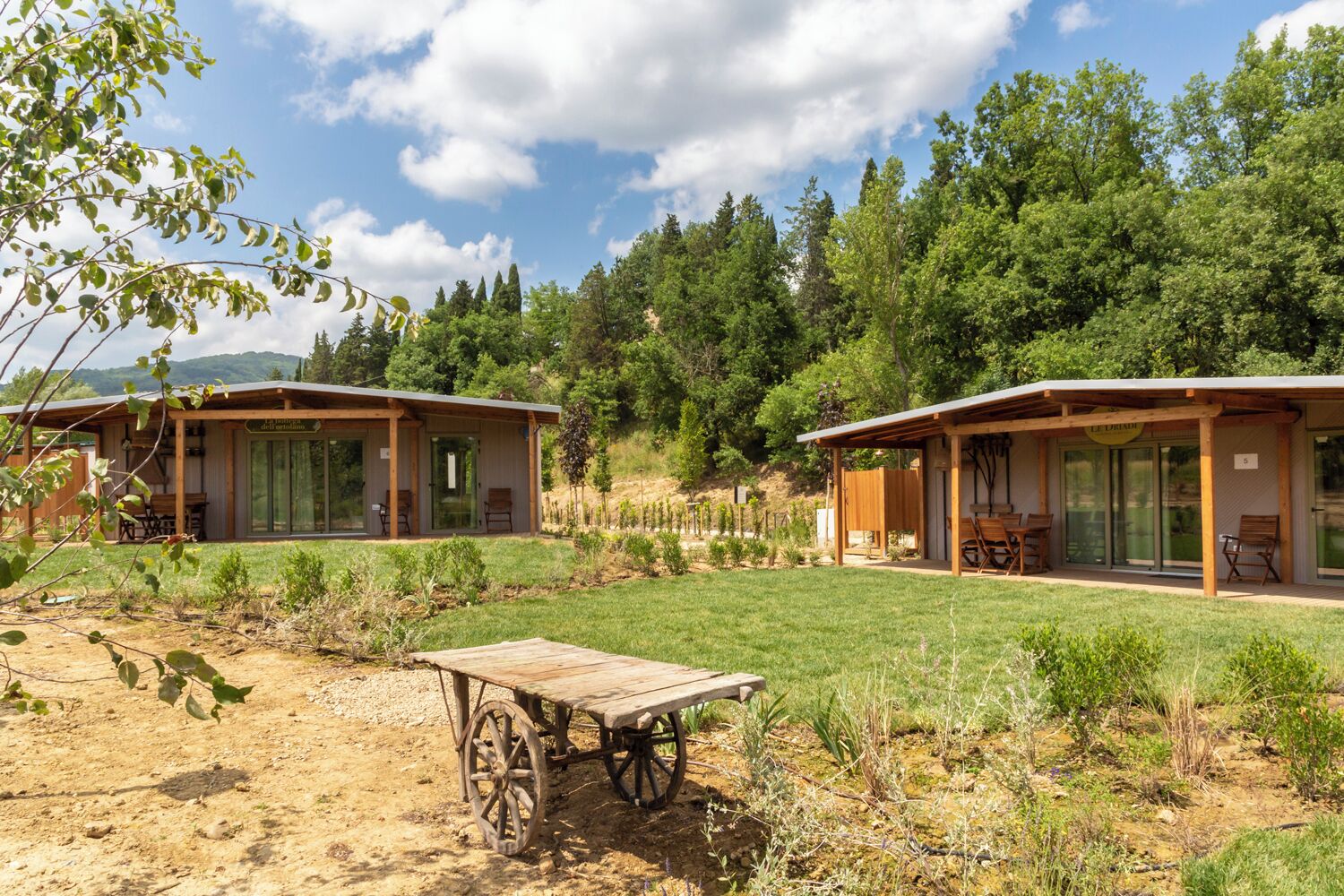 Deux cabanes en bois dotées de portes en verre, une charrette en bois et une zone herbeuse avec des arbres en arrière-plan.