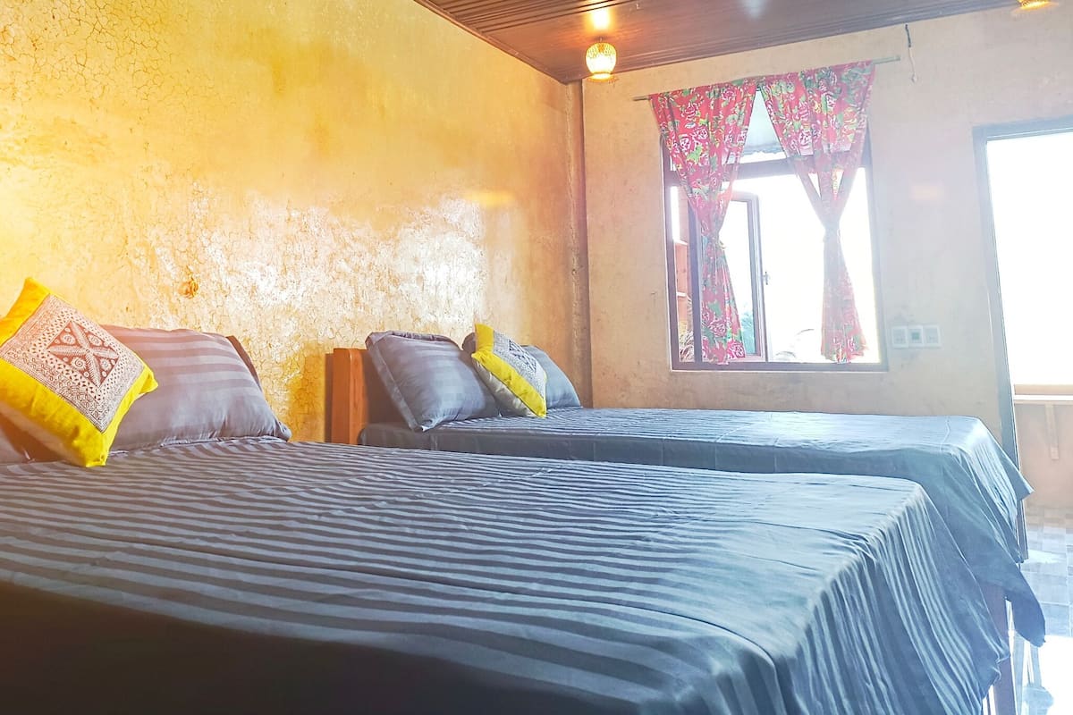 A room with two beds, a window with curtains, and a wooden ceiling.