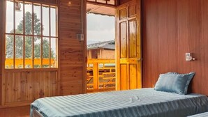 A wooden cabin room with a bed, a window, and a view of a wooden deck and trees. - Lý Gia Homestay (Quan Ba)