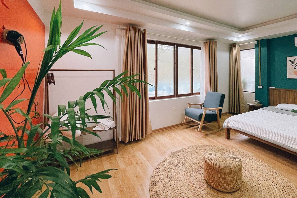 A room with a bed, a chair, a potted plant, a woven ottoman, and a window with curtains.