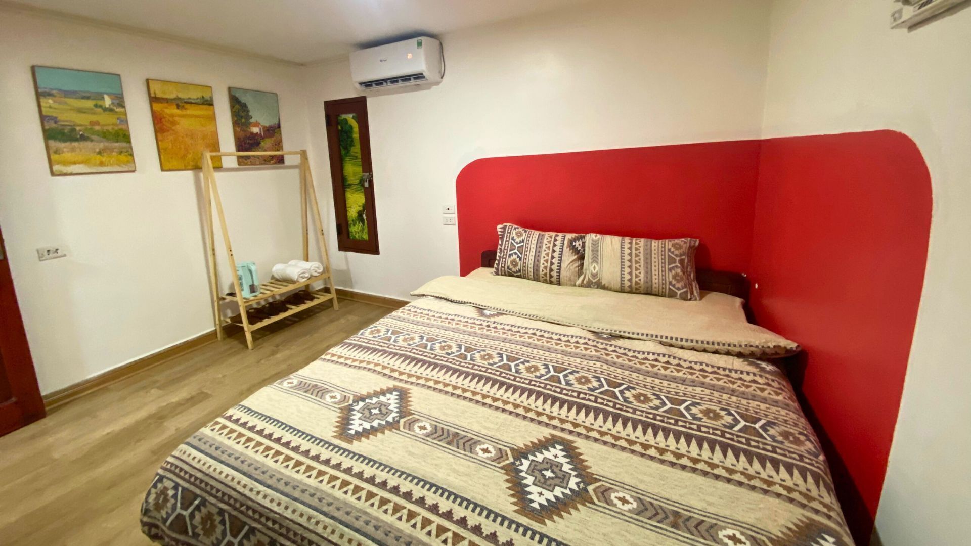 Un dormitorio con un cabecero rojo, una colcha estampada, un tendedero de madera y tres cuadros de paisajes en la pared.