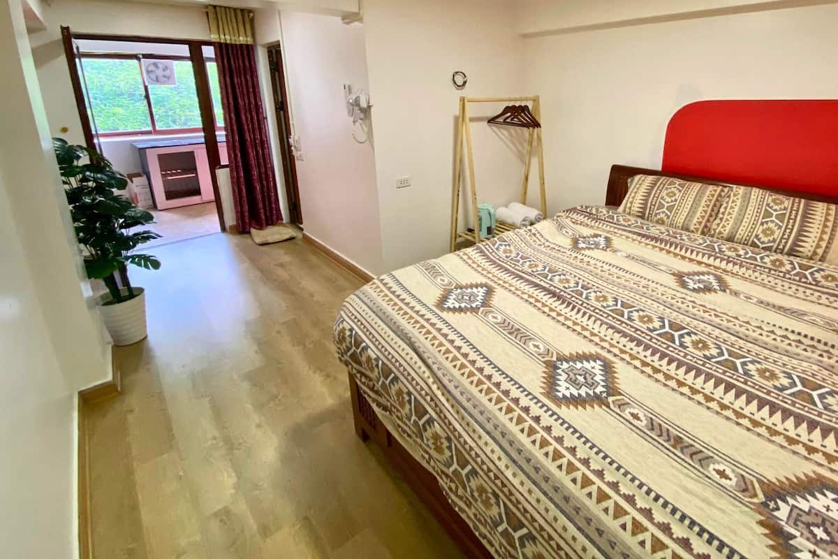 A bedroom with a patterned bedspread, a red headboard, a wooden bed frame, a potted plant, and a view of a balcony with curtains.