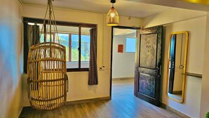 Un pasillo con una silla de mimbre, un candelabro colgante, una puerta con un número y un espejo en la pared.