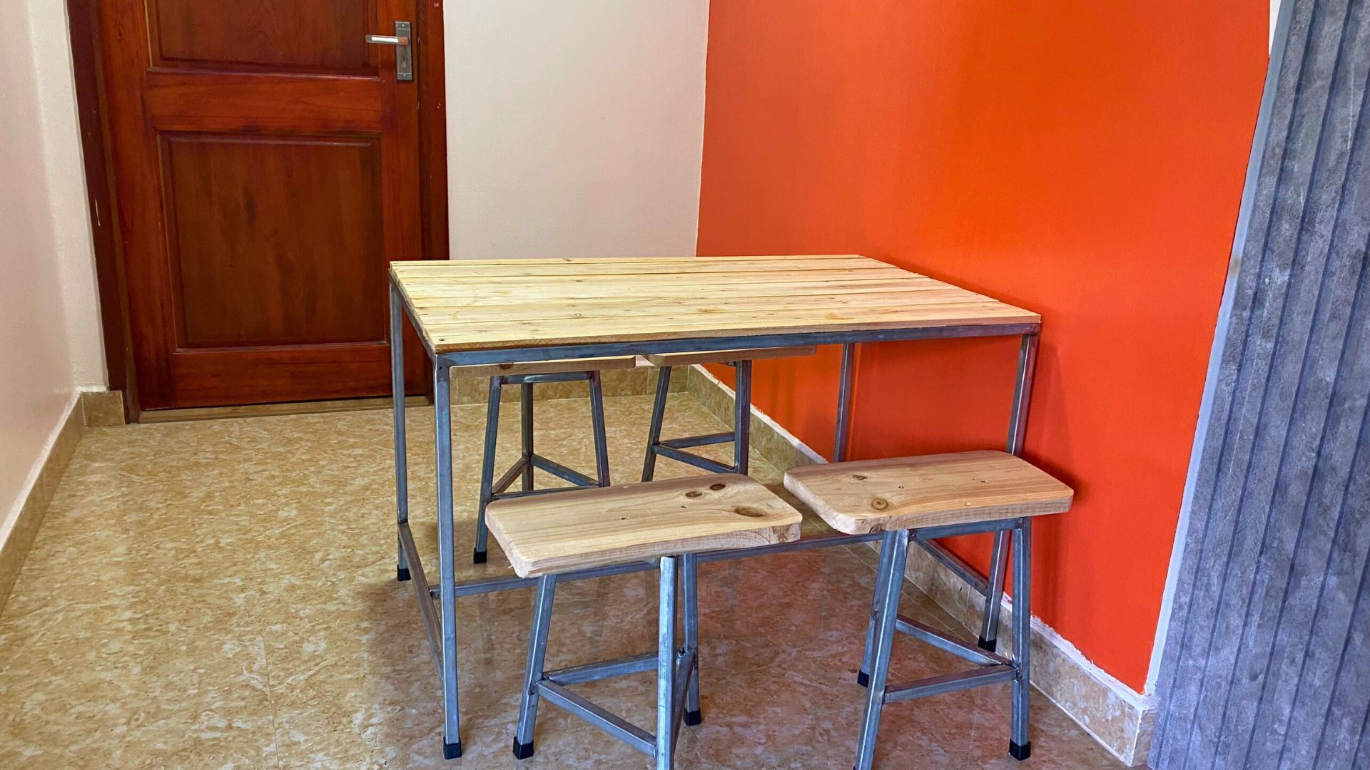 A small wooden table with metal legs and a bench attached to it, positioned against a red wall in a room with a wooden door and tiled floor.