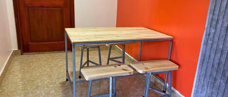 A small wooden table with metal legs and a bench attached to it, positioned against a red wall in a room with a wooden door and tiled floor.