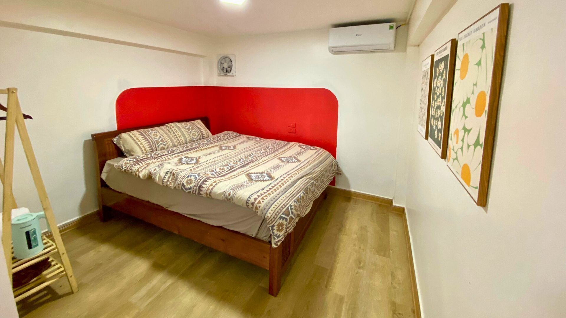 A small bedroom with a wooden bed, a red headboard, and a wooden floor. There is a small table with a chair and a ladder in the corner.