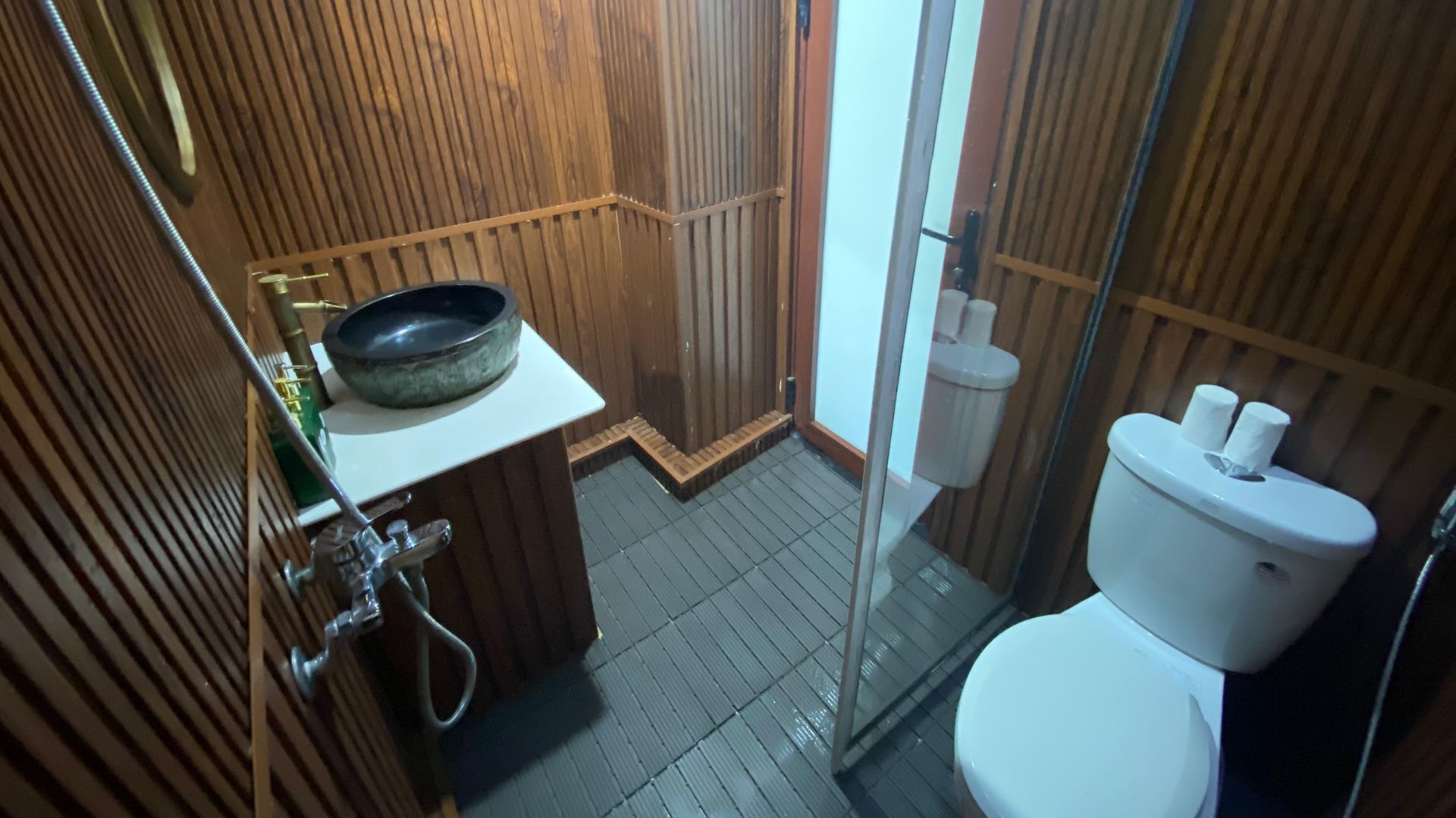 A small, compact bathroom with a wooden vanity, a glass shower enclosure, and a white toilet.