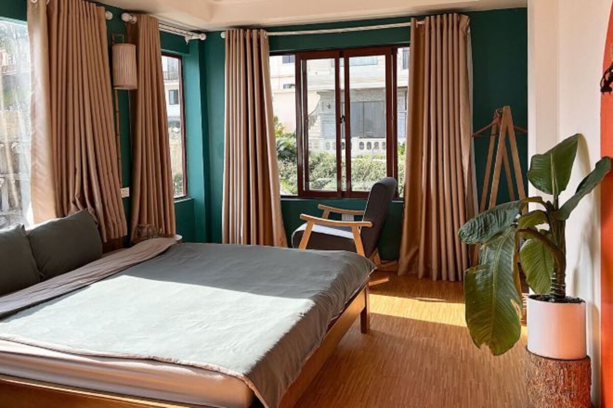 A bedroom with a bed, a desk, and a chair. There is a window with curtains and a view of buildings outside.