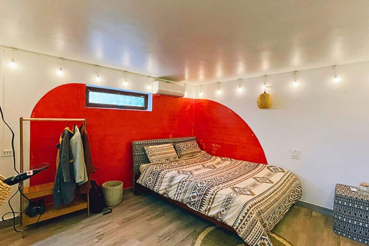A compact bedroom with a red accent wall, a bed with patterned bedding, a small wardrobe, and a hanging light fixture.