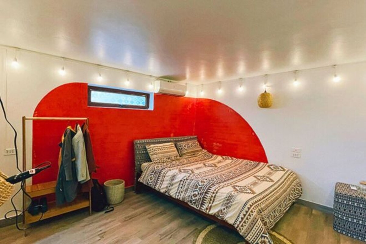 A compact bedroom with a red accent wall, a bed with patterned bedding, a small desk with a chair, and string lights on the ceiling.