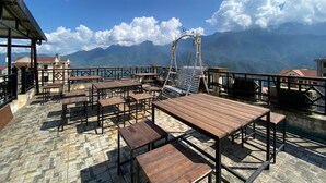 Una terraza en la azotea con mesas y sillas de madera, baranda metálica y vistas a montañas al fondo.
