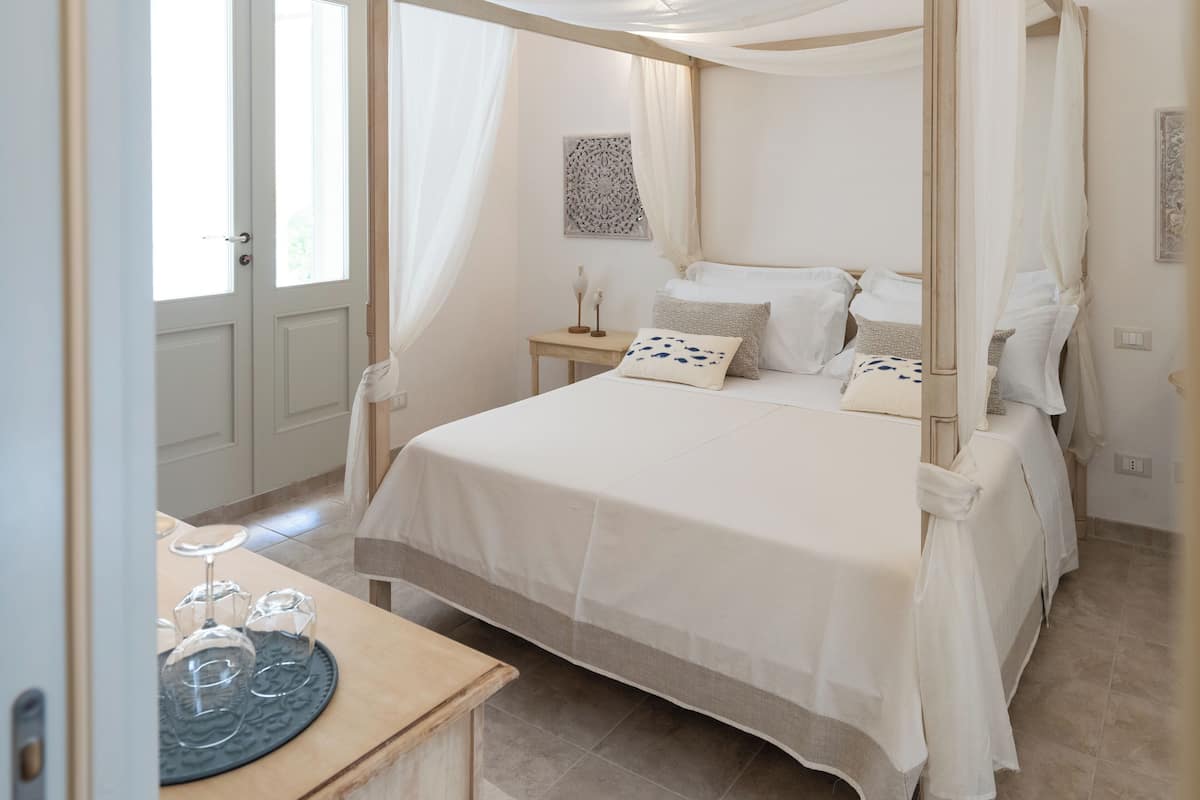 A four-poster canopy bed with white linens, a bedside table with candles, and a view of the outdoors through a door.