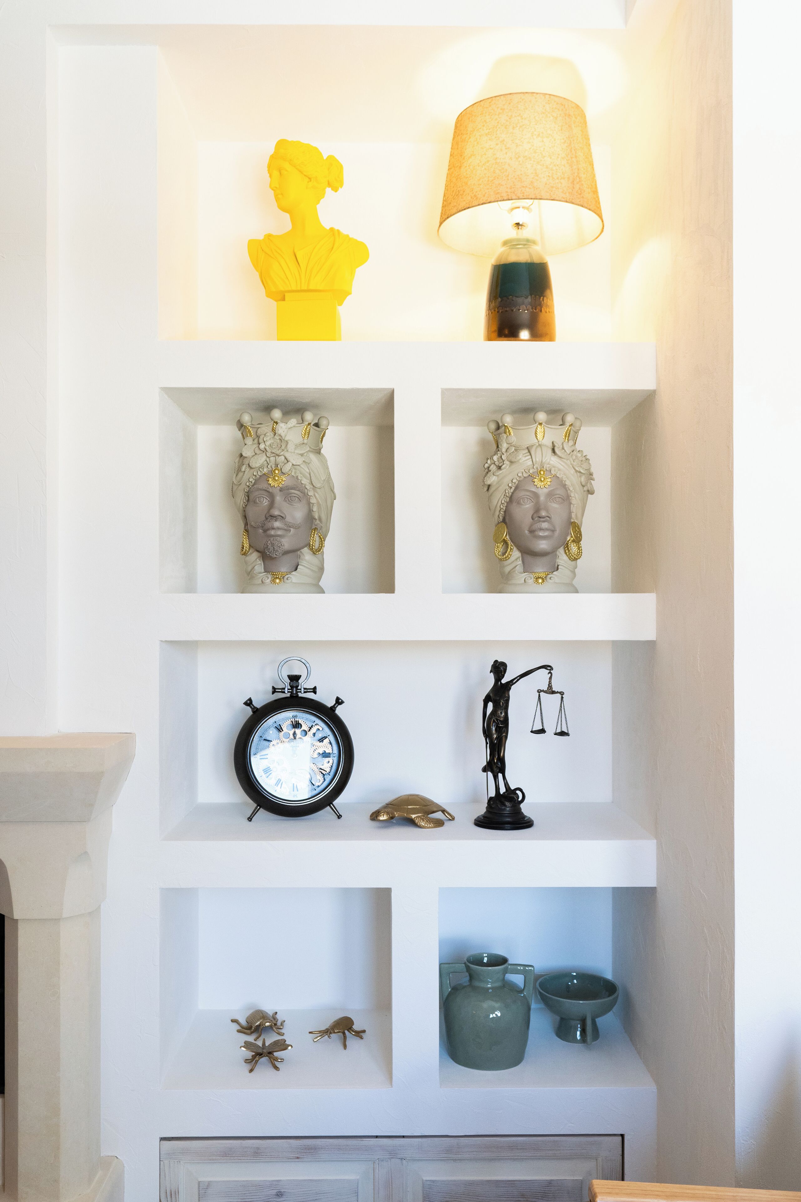 A white shelf with decorative items including a yellow bust, a lamp, two ornate busts, a clock, a scale figurine, and two ceramic vases.