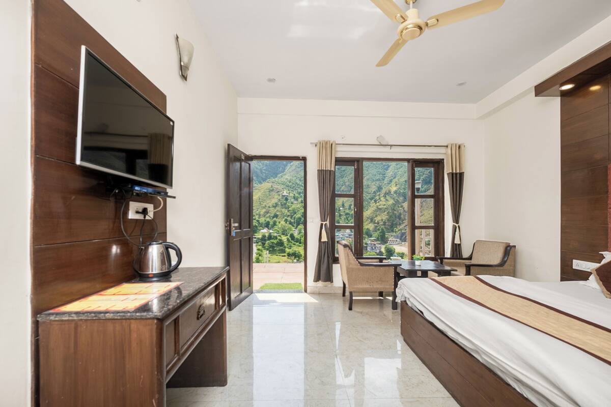 A hotel room with a flat-screen TV, a ceiling fan, a bed, a desk, a chair, and a view of a mountainous landscape through the window.