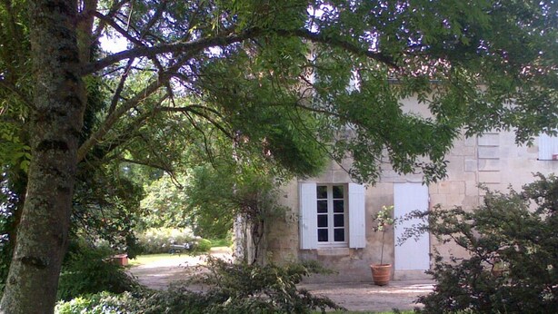 A stone building with a white door and windows, surrounded by lush greenery and a tree in the foreground. - A haven of tranquillity on the banks of the Dordogne River (Fronsac)