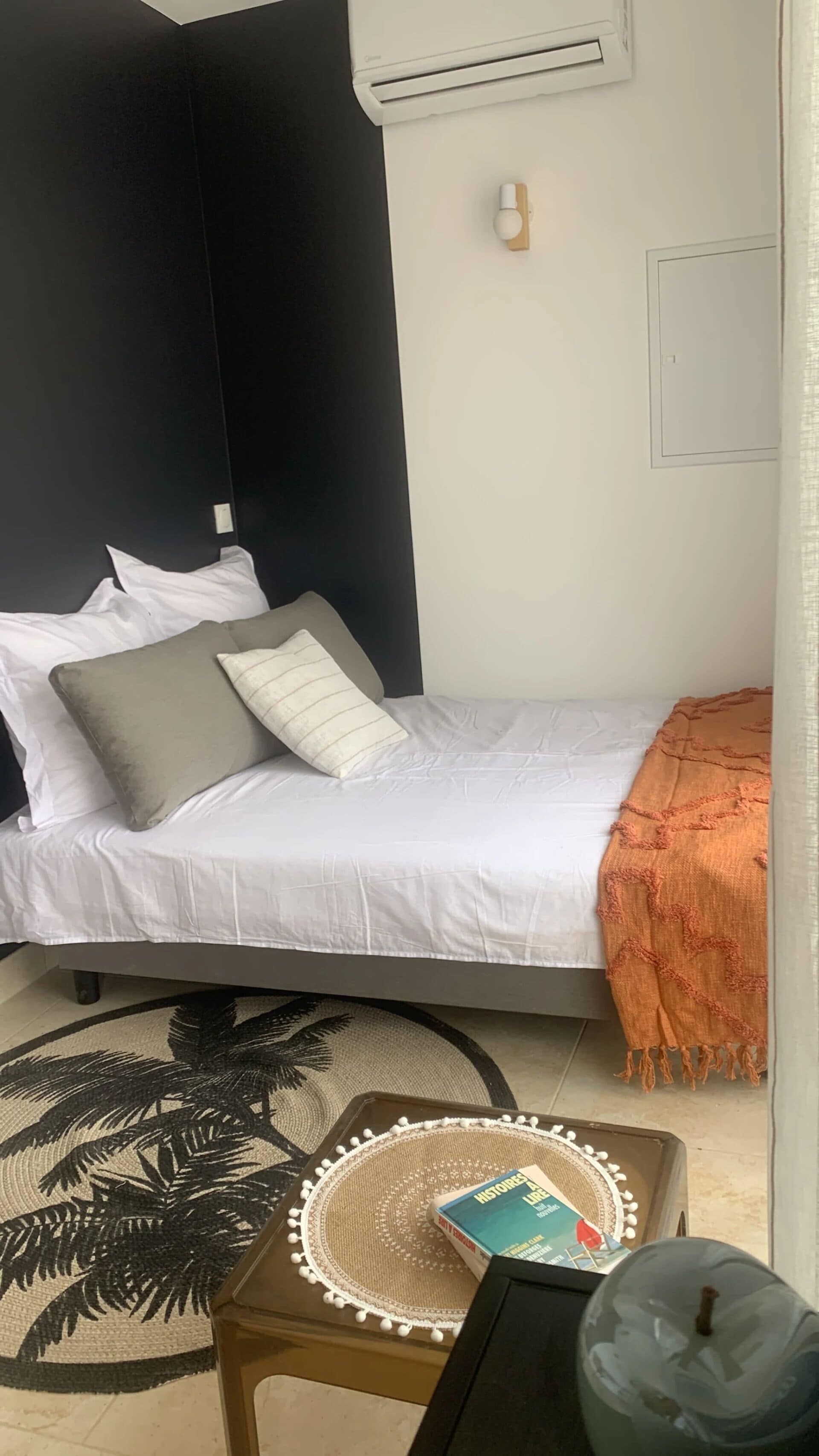 A bedroom with a bed, pillows, a blanket, a small table with a book, a decorative mat, and a wall-mounted air conditioner.