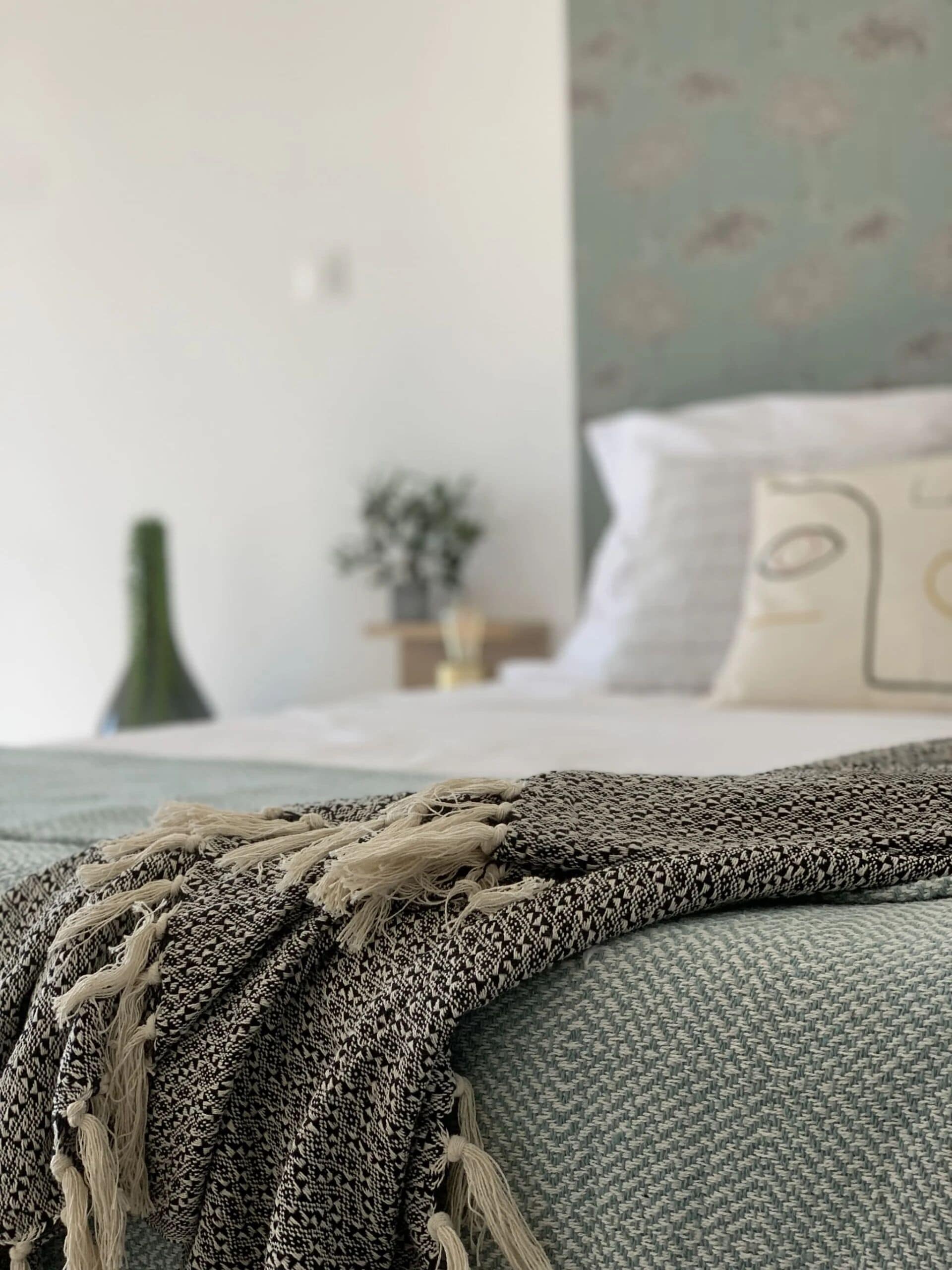 Un lit recouvert d’une couverture à motifs gris et blancs, un oreiller vert et un mur orné d’un papier peint à motifs floraux discrets.
