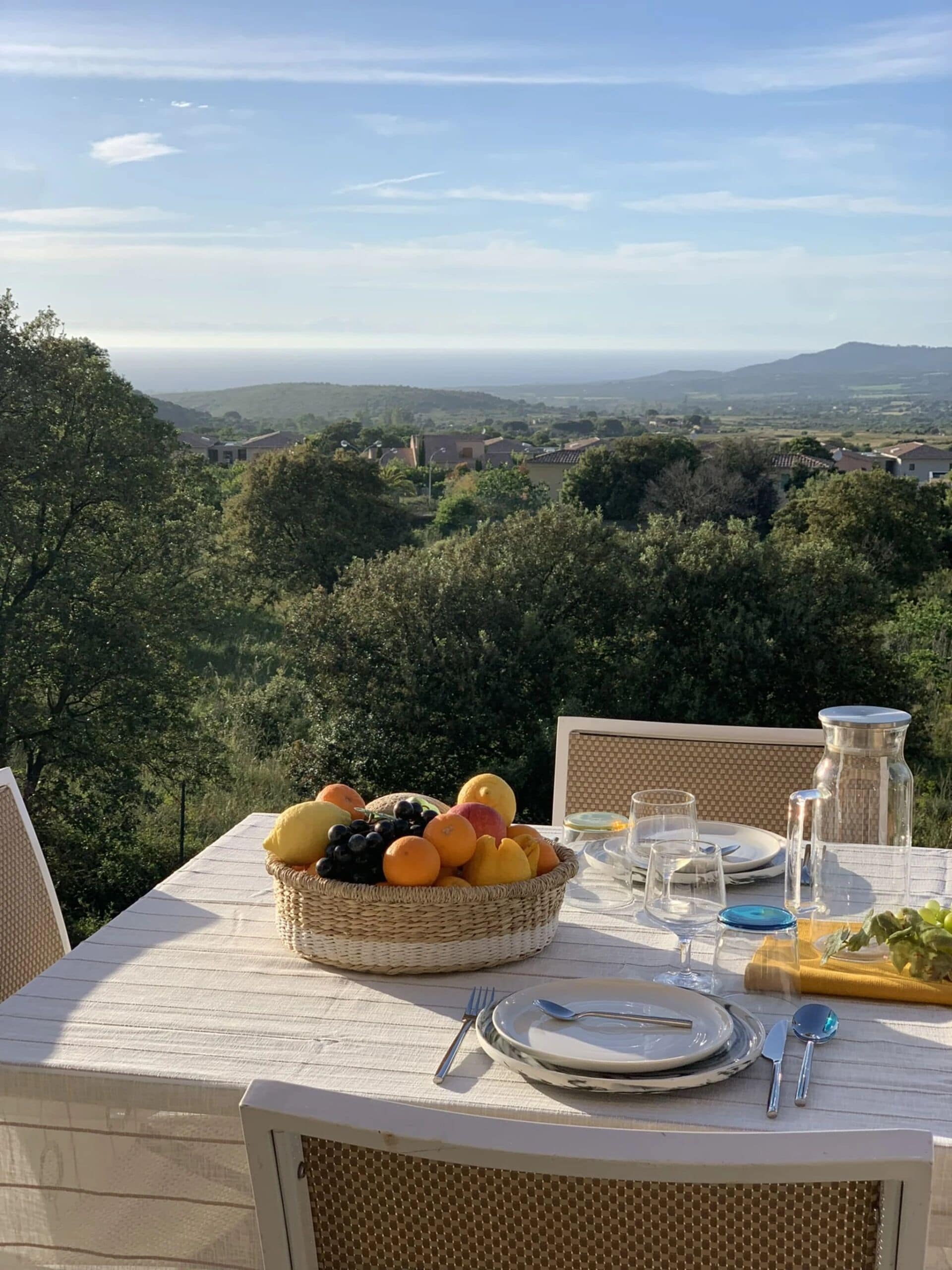 Une table dressée pour un repas, avec un panier de fruits, des assiettes, des couverts et des verres, donnant sur un paysage pittoresque avec des arbres et des collines au loin.